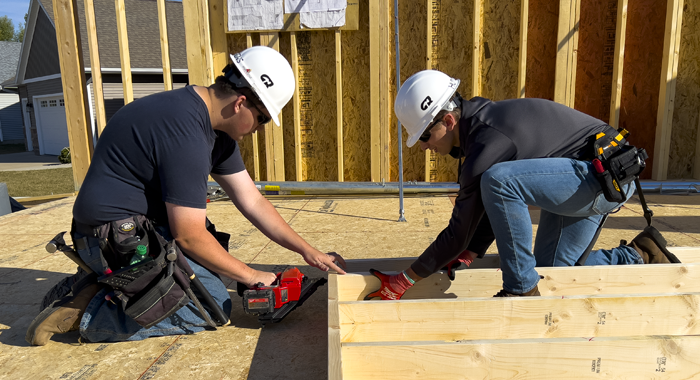 Two crew members of the student built house use an air nailer to frame a wall.
