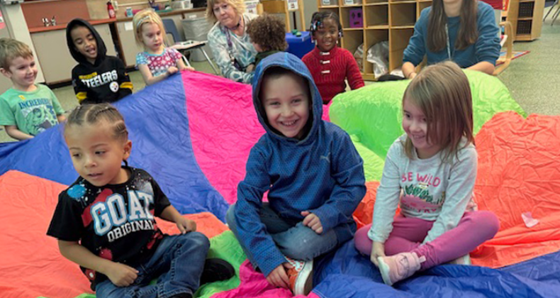 Preschool students sit on a parachute during a gym class.